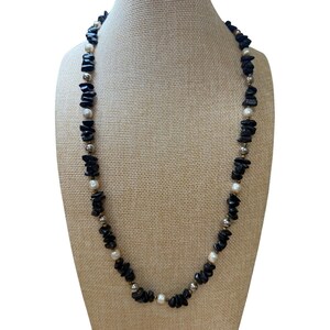 May include: A necklace featuring a repeating pattern of irregular, tumbled black stone chips interspersed with smooth, round white pearls and small, polished silver-toned metallic beads, arranged in a single strand on a flexible cord.