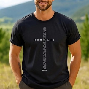 Könnte beinhalten: Ein schwarzes Kurzarm-T-Shirt mit einem weißen Grafikdesign auf der Brust. Das Design zeigt das Wort ROMANS mit einem vertikalen Kreuz, das aus dem Text des Bibelverses Römer 8,28-29 gebildet wird, inklusive der Quellenangabe am unteren Rand.