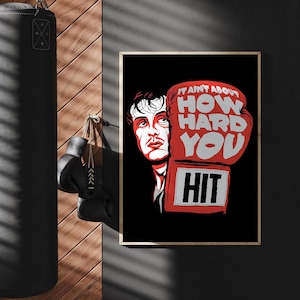 May include: A black punching bag hangs next to a framed poster with a boxing theme. The poster features a portrait of a man with a red boxing glove that reads "IT AIN'T ABOUT HOW HARD YOU HIT."