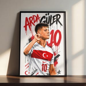 May include: Framed art print featuring a stylized portrait of Arda Güler, a soccer player. He is wearing a white and red jersey with the number 10 and the Turkish flag. The artwork includes the player's name and number in bold, graffiti-style lettering.