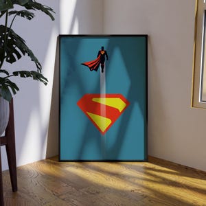 May include: Framed poster with a minimalist Superman design. The poster has a teal background with a large red and yellow Superman logo at the bottom. A silhouette of Superman in flight is at the top, with a red cape.