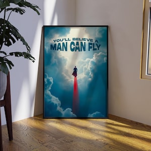 May include: Framed poster featuring a superhero soaring through a cloudy sky. The poster has the text "YOU'LL BELIEVE A MAN CAN FLY" in white and blue. The image is set in a corner with a plant and wooden floor.