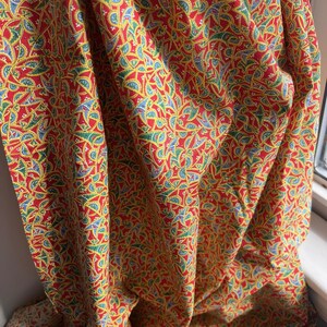 May include: Red fabric with a repeating pattern of yellow, blue, and green shapes. The fabric is draped, showing folds and texture. The design is intricate and detailed, creating a visually rich textile.