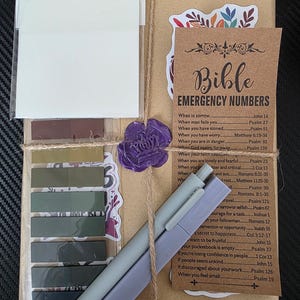 Può includere: Un set regalo avvolto in carta marrone, legato con spago e sigillato con un sigillo di cera viola. Include un blocco note, note adesive, una scheda dei numeri di emergenza della Bibbia e due penne. Il set è decorato con adesivi floreali e linguette adesive.