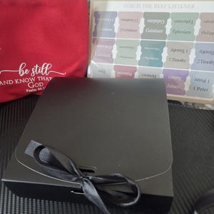 May include: A red canvas pouch with the white text 'be still AND KNOW THAT GOD Psalm 46:10', a black gift box tied with a black ribbon, and a clear plastic sheet containing colorful adhesive Bible index tabs labeled 'GOD IS THE BEST LISTENER'.