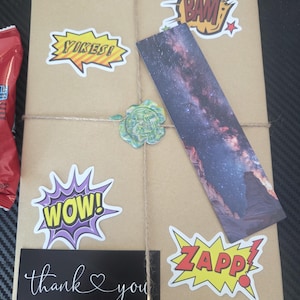 May include: A wrapped package in brown paper, tied with twine, and decorated with comic book-style stickers that say "YIKES!", "BAM!", "WOW!", and "ZAPP!". A bookmark with a galaxy image and a thank you card are also attached.