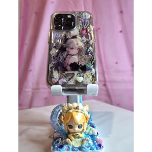 May include: Clear phone case with a decorative anime-style illustration of a woman with long white hair and a black dress. The case is on a white phone stand with a figurine of a blonde anime character.
