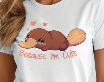 May include: A white short-sleeved t-shirt featuring a graphic of a brown platypus with large blue eyes, two small pink hearts above it, and the text "Because I'm Cute" printed in a light orange script font across the center of the chest.