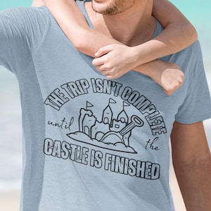 Op de afbeelding: Een lichtblauw T-shirt met een zwarte afbeelding waarop staat "De reis is pas compleet als het kasteel klaar is". De afbeelding toont een zandkasteel met vlaggen, een emmer en een schep.