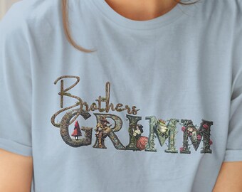 Może przedstawiać: Jasno błękitny t-shirt z kwiatowym wzorem, który tworzy napis "Brothers Grimm".