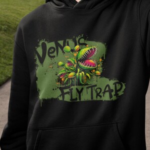 Puede incluir: Sudadera con capucha negra con un gráfico verde de plantas de Venus atrapamoscas. El gráfico tiene el texto "Venus Fly Trap" en blanco.