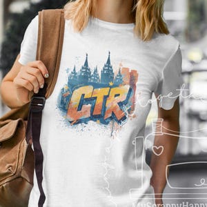 Op de afbeelding: Wit T-shirt met een kleurrijk grafisch ontwerp met de letters "CTR" in oranje en blauw, en een silhouet van een stadsgezicht. Het shirt wordt gedragen door een persoon met een zonnebril en een bruine rugzak.