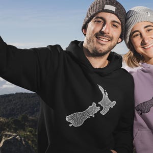 This image features a smiling couple taking a selfie on top of a mountain, wearing hoodies.  The design on the front of the pullover jackets is either white or black outline of the New Zealand country filled with intricate Maori tattoo inspired designs.
