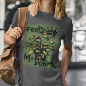 Puede incluir: Una camiseta gris oscuro con un gráfico verde de una planta carnívora con el texto "Feed Me Power" y una corona.