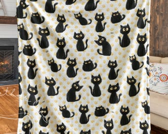 May include: A white fleece blanket featuring a repeating pattern of black cat illustrations and yellow paw prints. The cats have yellow eyes and are in various poses. The blanket is held up by a person.