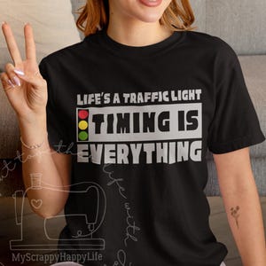 Puede incluir: Camiseta negra con el texto blanco "LIFE'S A TRAFFIC LIGHT, TIMING IS EVERYTHING". El diseño incluye un semáforo. Es una prenda informal.