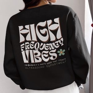 Op de afbeelding: Zwart sweatshirt met een witte grafische print die "HIGH FREQUENCY VIBES" zegt met een kleine bloemgrafiek. De tekst bevat ook "IT'S WHATS INSIDE THAT COUNTS" en "ULTRASOUND DEPARTMENT".