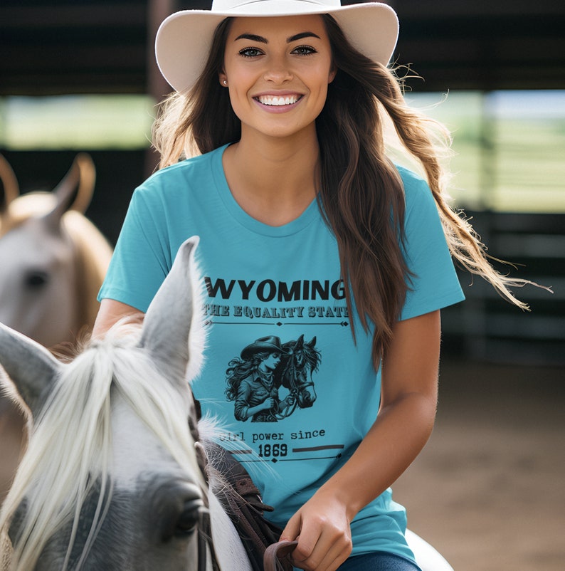 May include: A woman wearing a turquoise blue t-shirt with a graphic of a cowgirl riding a horse and the text "Wyoming, The Equality State, Girl power since 1869". She is wearing a white cowboy hat and is sitting on a horse.