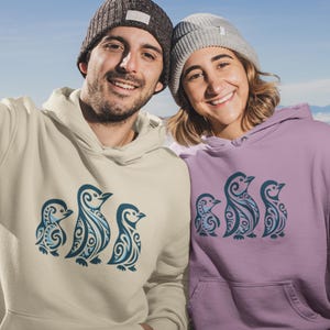 May include: Two people wearing hoodies with a design of stylized penguins. The design features three penguins with intricate patterns. The hoodie on the left is cream colored and the hoodie on the right is lavender.