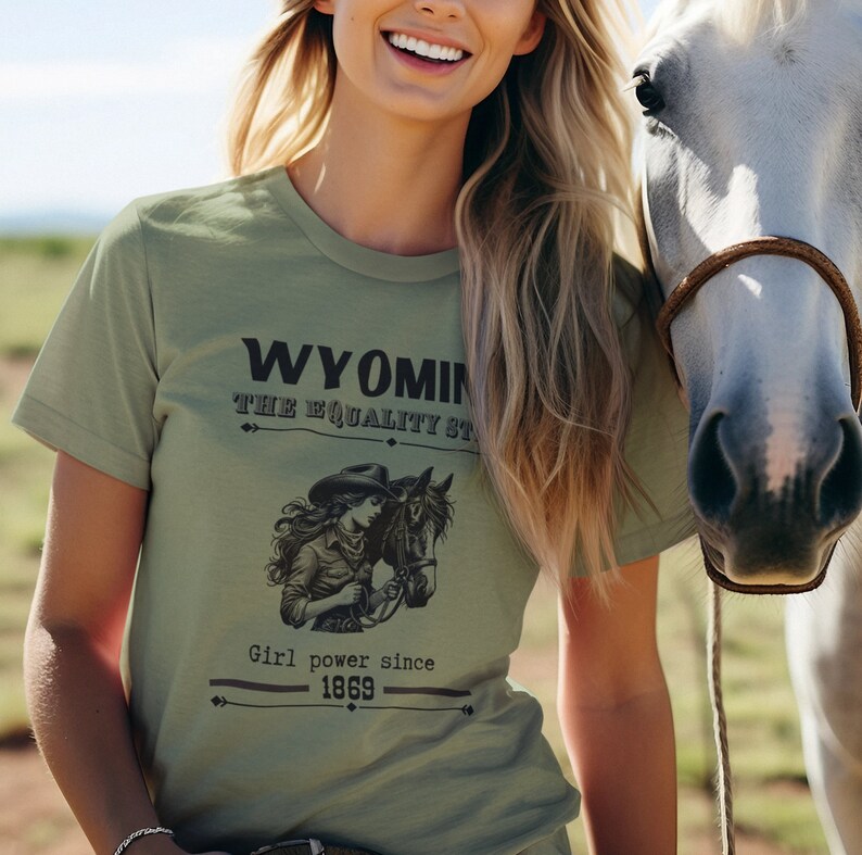 May include: A woman wearing a green t-shirt with a graphic design that says "Wyoming The Equality State" and "Girl power since 1869". She is standing next to a white horse.