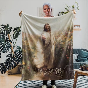 May include: A beige blanket featuring a religious image of Jesus extending his hand. The words "Walk With Me" are written in cursive script across the bottom. The blanket is held up by a person.
