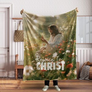 May include: A soft throw blanket featuring a printed image of a person with long hair sitting in a field of colourful wildflowers and tall grass, holding a butterfly. The text "Rejoice In CHRIST" is printed in large white letters at the bottom.