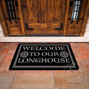 Puede incluir: Una alfombra rectangular negra con las palabras "WELCOME TO OUR LONGHOUSE" en blanco, enmarcada por un borde decorativo. La alfombra está colocada frente a una puerta de madera.