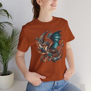 May include: A rust-colored t-shirt with a graphic of a blue and green dragon with wings spread, sitting on a crescent moon with feathers and beads.