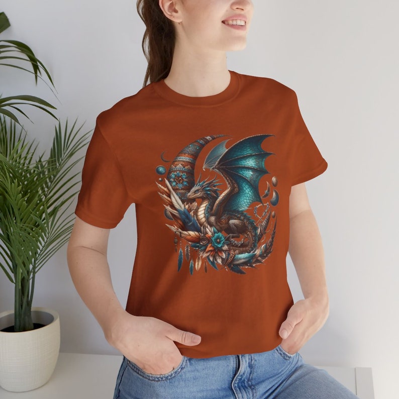 May include: A rust-colored t-shirt with a graphic of a blue and green dragon with wings spread, sitting on a crescent moon with feathers and beads.