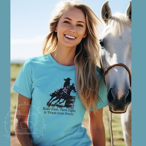 Op de afbeelding: Een lichtblauw t-shirt met een zwarte afbeelding van een paard en ruiter in een barrel racing scène. De tekst "Ride Fast, Turn Tight & Trust your Horse" staat onder de afbeelding. De persoon die het t-shirt draagt heeft lang blond haar.