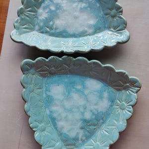 May include: Two turquoise ceramic plates with a scalloped edge and a floral design. The plates have a triangular shape with a light blue interior and a darker blue border. The plates are handmade and have a rustic, artisanal feel.