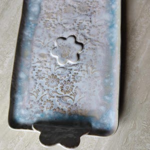 May include: Rectangular ceramic tray with a floral pattern and a flower-shaped cutout. The tray has a pearlescent finish with blue and gold accents. The tray has a handle on each end.