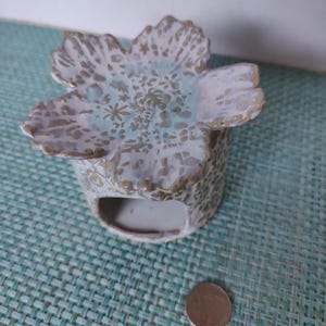 Pode incluir: Um queimador de incenso de cerâmica branca em forma de flor com um furo no centro. O queimador tem um design floral e está sentado em um tapete tecido verde e branco.