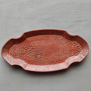 Op de afbeelding: Een ovaalvormige serveerschaal van terracotta-kleurig keramiek. De schaal heeft een reliëf bloemen- en vlinderontwerp. De randen van de schaal hebben een geschulpte vormgeving. Deze decoratieve schaal is geschikt voor het serveren van voedsel of het presenteren van items.