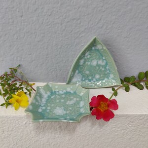 May include: Two ceramic dishes with a turquoise glaze and white crystalline patterns. One dish is triangular, the other is rectangular with scalloped edges. The dishes are displayed on a white surface with flowers.