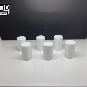 May include: Six white cylindrical objects with rounded tops are arranged on a white surface. The objects are evenly spaced and appear to be small, possibly bottle stoppers or similar components. The background is a gradient of black and white.