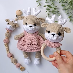 Könnte beinhalten: Handgefertigtes Häkeltier-Spielzeug und Zubehör in Beige, Weiß und Rosa. Das Set enthält eine Rehpuppe mit rosa Kleid, eine Rassel und einen Schnullerhalter mit Holzperlen. Die Rehe haben weiße Geweihe und schwarze Augen.