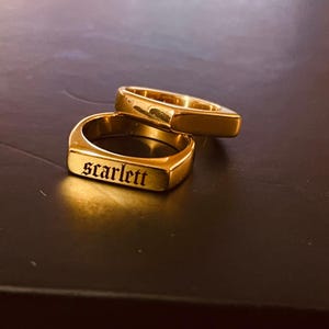 Peut inclure: Deux chevalières dorées. La bague avant porte le nom "Scarlett" gravé en police gothique. Les bagues sont rectangulaires avec des bords arrondis et sont présentées sur une surface sombre.