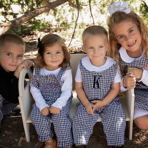 May include: Four children wearing black and white gingham overalls and shirts. The children are sitting on a white bench and smiling at the camera. The girl on the right is wearing a white bow in her hair.