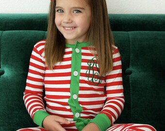 May include: A young girl wearing a red and white striped long-sleeved pajama onesie with green trim and a green monogram on the chest. She is sitting on a green couch with a red bow in her hair.