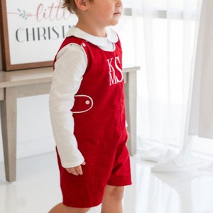 May include: A young child wearing a red short-sleeved overall with white piping and a white long-sleeved shirt. The overall has a white embroidered monogram "KSM".