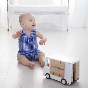 May include: A baby boy wearing a blue and white gingham romper with the name "Ricky" embroidered on the front, sitting on a white wood floor with a white toy truck with wooden details.