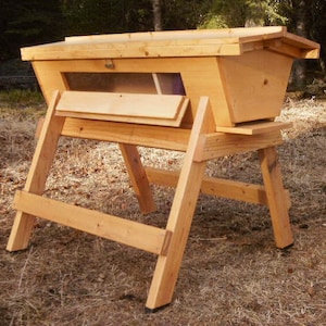 Puede incluir: Una colmena de madera con un techo inclinado y una sección inferior extraíble. La colmena está soportada por un marco de madera con dos patas a cada lado.