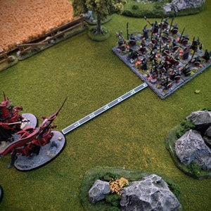 Op de afbeelding: Een tabletop wargaming-sc&egrave;ne met een zwarte plastic bewegingsmeter gemarkeerd met de cijfers &eacute;&eacute;n tot en met zes, geplaatst tussen een groep rood gepantserde bereden miniaturen en een vierkante formatie van donker geklede infanterie.