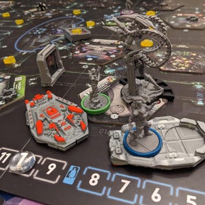 Op de afbeelding: Een bordspelopstelling met verschillende miniatuurfiguren en spelstukken op een donker spelbord. De stukken omvatten een grijs ruimteschipachtig figuur met rode accenten, een hoog, veelpotig wezen en kleine figuren op ronde bases.