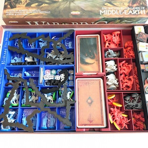 Pode incluir: Uma caixa de jogo de tabuleiro aberta, revelando compartimentos organizados cheios de peças do jogo. Os compartimentos são azuis e vermelhos, contendo miniaturas, cartas, dados e outros componentes do jogo. A tampa da caixa exibe o título do jogo "Epic Battles in the Lands of Middle-Earth".