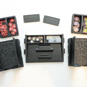 May include: Black 3D-printed board game organizer with multiple compartments and removable lids. Includes trays for tokens and cards. Features decorative side panels. Contains red, brown, and white game pieces.