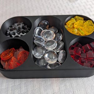 May include: A black plastic organizer with six compartments, each filled with different game pieces. The pieces include clear glass stones, red and yellow translucent shapes, and gray gear-shaped pieces. Ideal for board game organization.