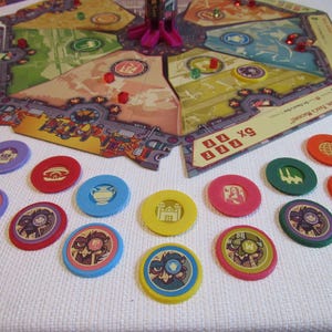 May include: A board game featuring a hexagonal board with colourful sections, a central pink structure, and small red and green cubes. Scattered in front are numerous circular tokens in various colours, each displaying unique graphic illustrations.
