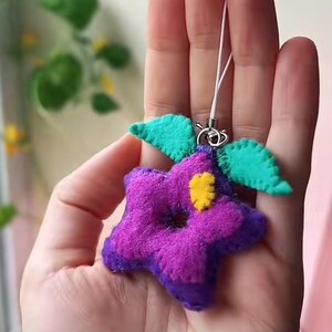 Pu&ograve; includere: Un fiore di feltro viola e blu con un centro giallo e foglie di feltro verde attaccate a una catena di ciondolo per telefono bianca.